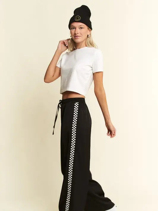 A woman modeling a white short-sleeve crop top and black wide-leg pants with a white-and-black checkered side stripe, paired with a black beanie, against a plain cream background.