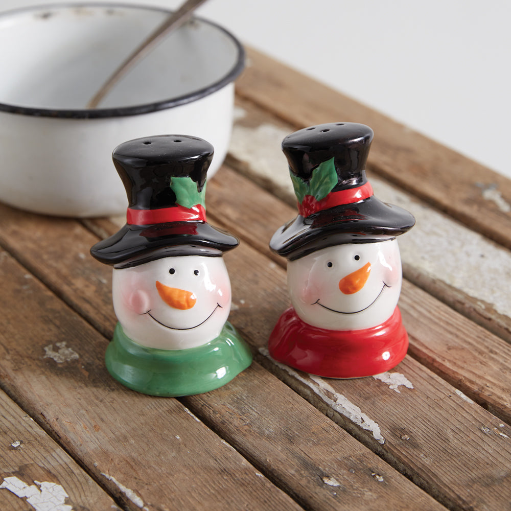Two snowman-themed salt and pepper shakers on a wooden surface with a white bowl in the background.