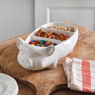 White pig-shaped bowl with compartments filled with snacks on a wooden surface.