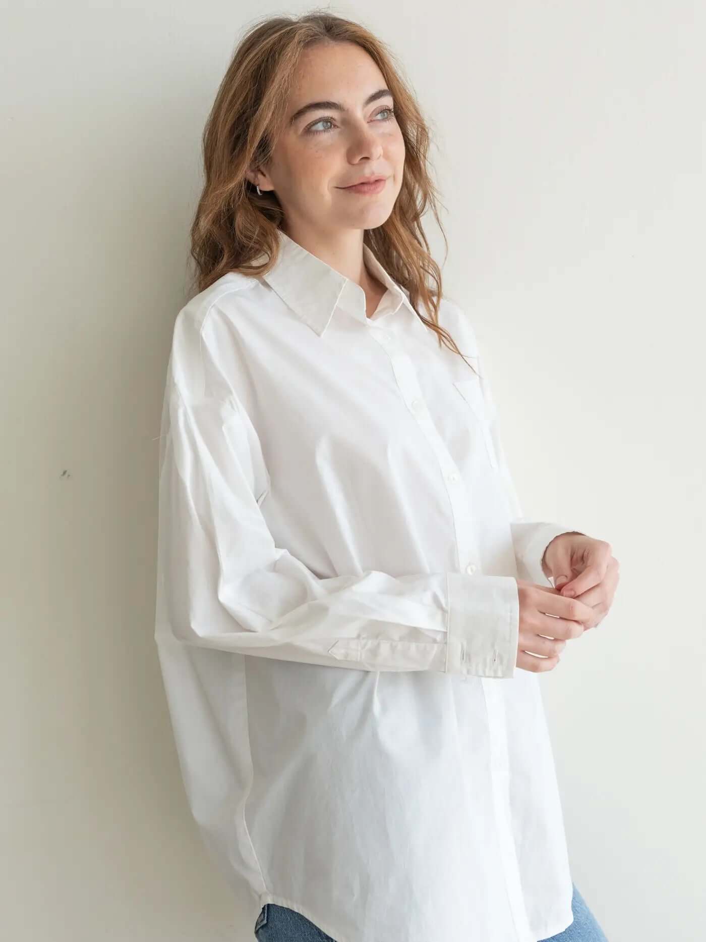 Woman in a white shirt and blue jeans standing in a bright room with a vase of flowers on a small table.