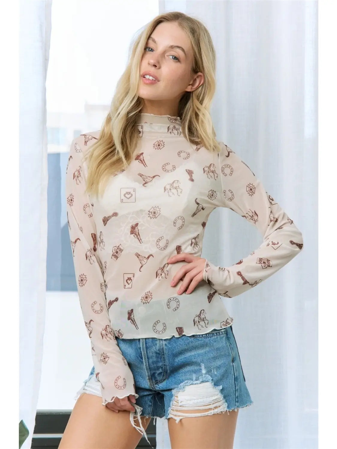 A blonde woman wearing a sheer long-sleeve mock neck top with a brown graphic pattern, paired with distressed denim shorts, posing in front of white curtains.
