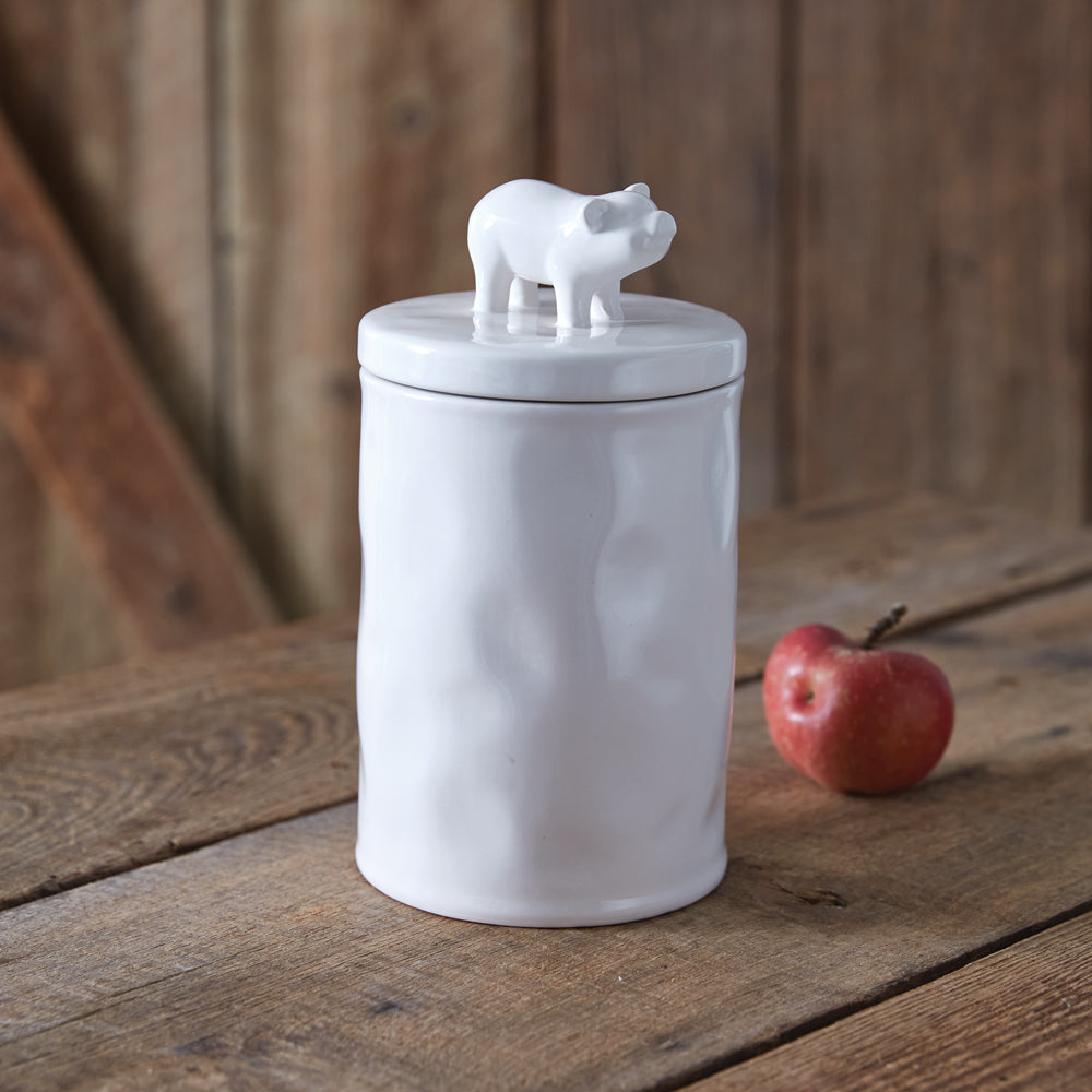 White ceramic jar with a piglet on top, placed on a wooden surface with an apple beside it.