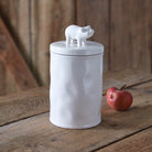 White ceramic jar with a piglet on top, placed on a wooden surface with an apple beside it.