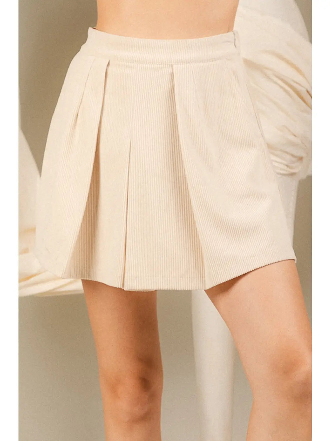 A close-up of a model wearing a pleated corduroy mini skirt in a light cream or beige color.