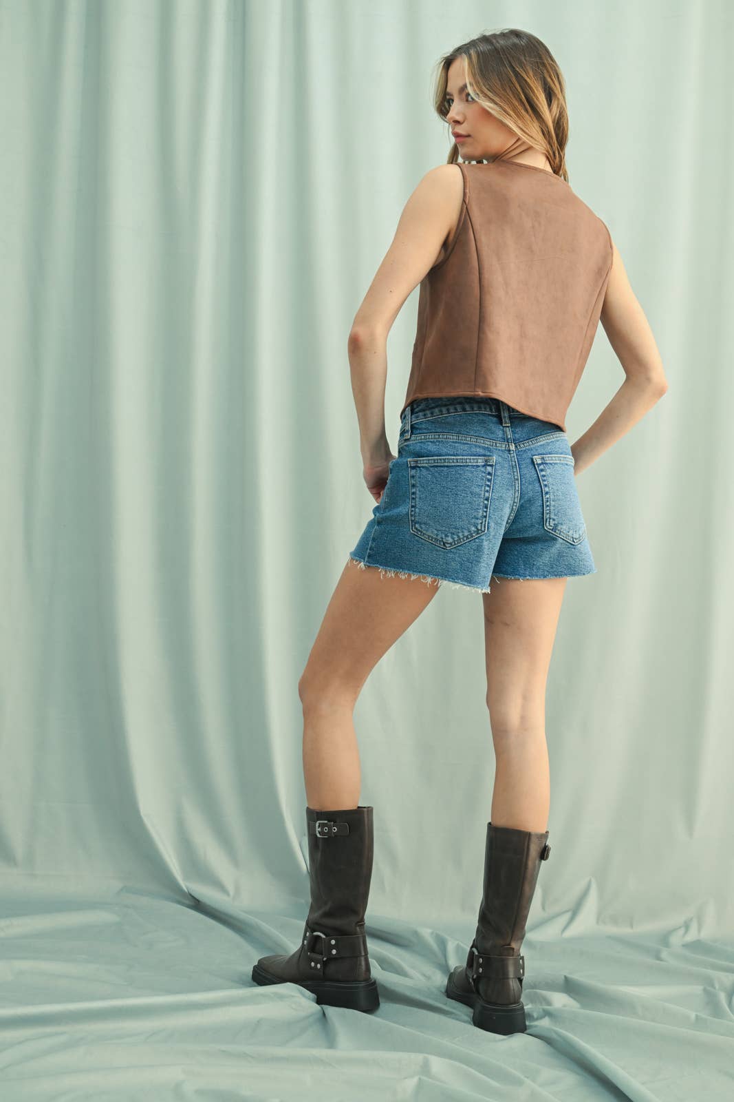 Back of Aa woman wearing a camel suede button-up vest over a white shirt, paired with Denim Shorts