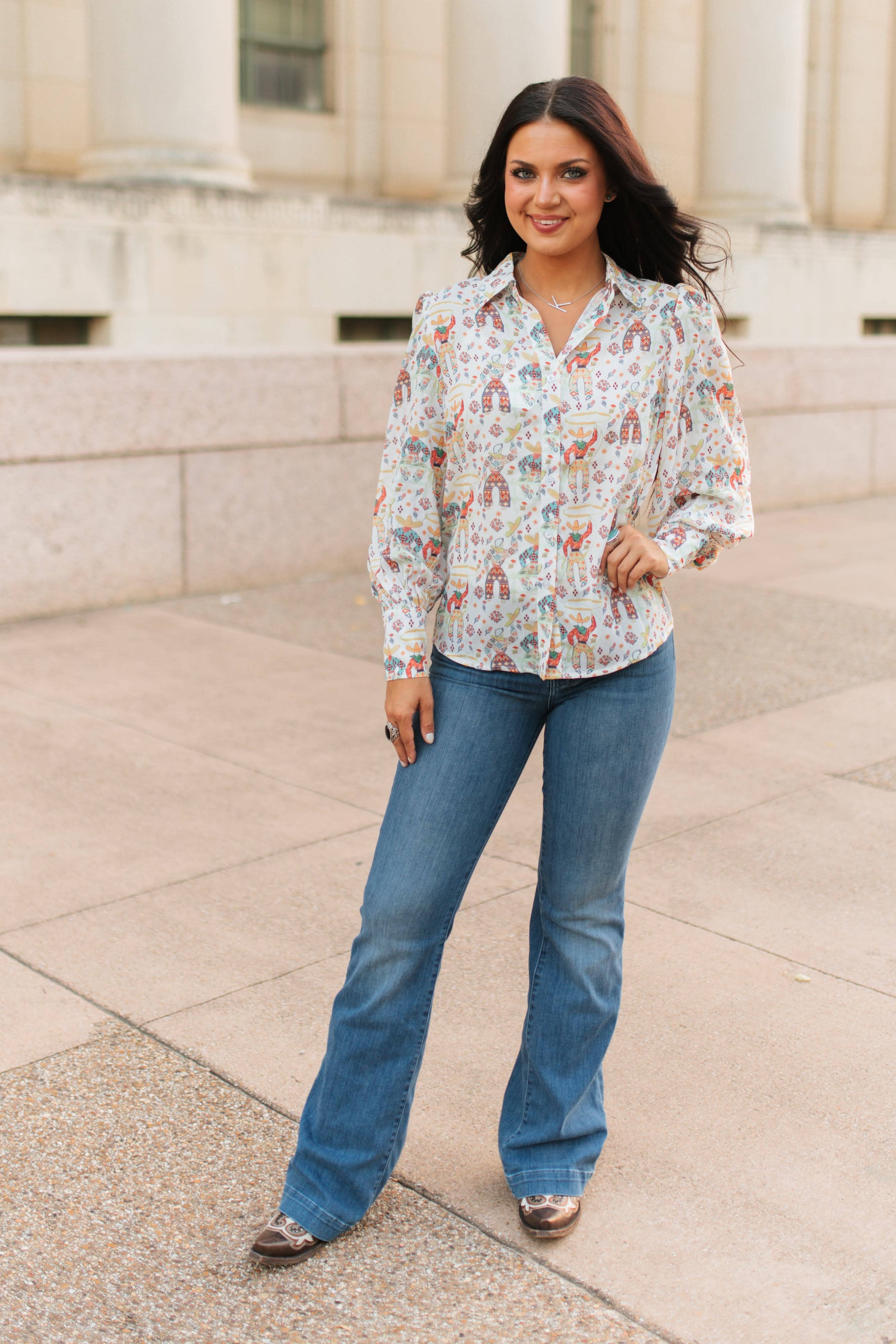 Woman wearing a patterned blouse and blue jeans standing on a stone pavement.