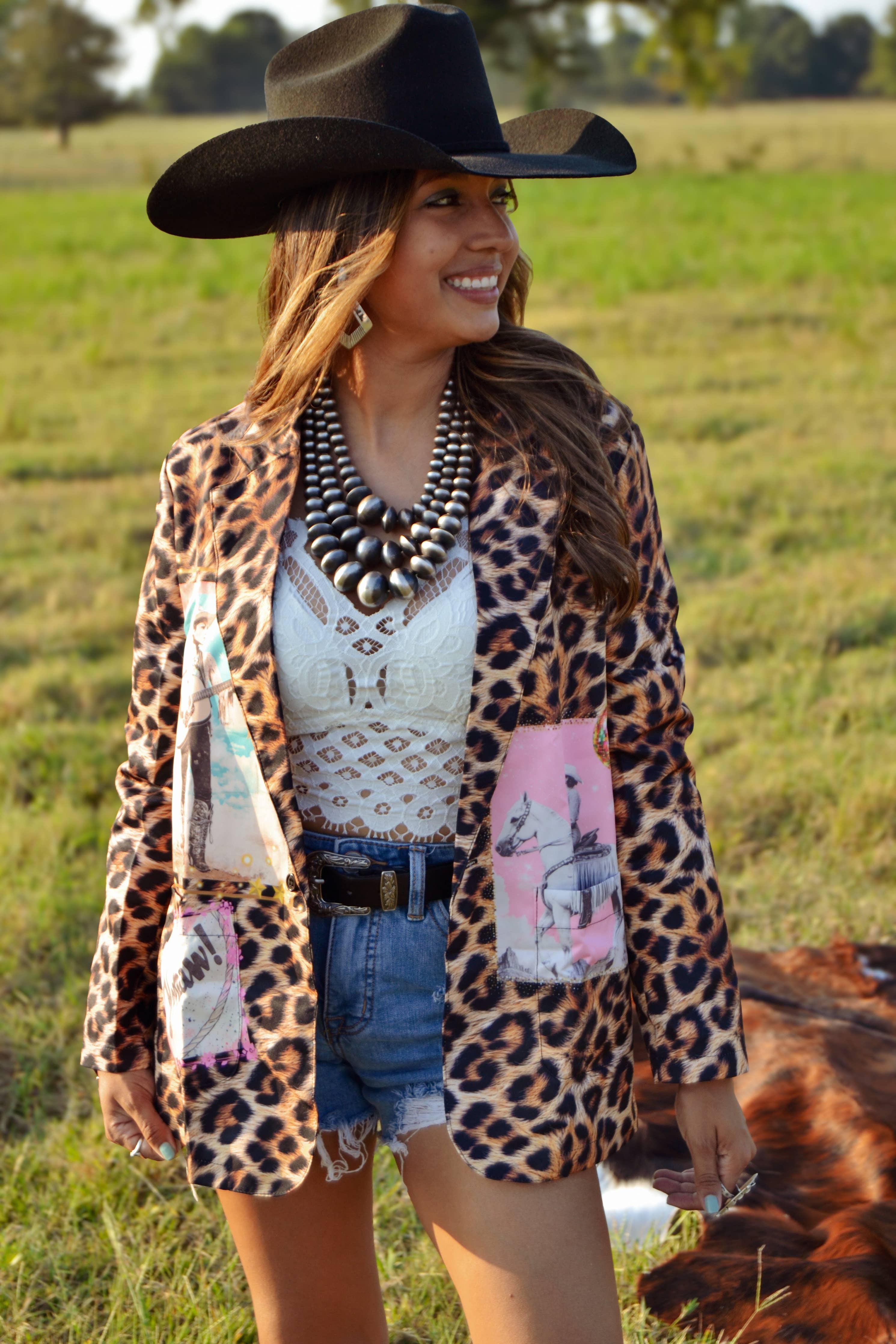 Woman in a leopard print cardigan and cowboy hat standing in a field with a cow.