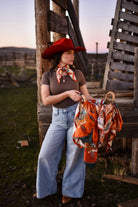 Woman in cowboy hat and scarf holding a patterned bag in a rustic outdoor setting