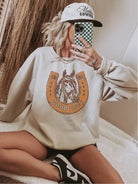 Person wearing a beige sweatshirt with a horse graphic and text, holding a phone with a checkered case.