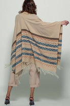 Woman wearing a beige poncho with a  Tan background and light blue, brown, light rust, and grey aztec pattern design  in stripes with tassel trim along bottom hem. Light weight fabric Full back view