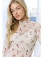 A blonde woman wearing a sheer, long-sleeve cream top with a brown western-themed print of horseshoes, lasso, boots, and cards.