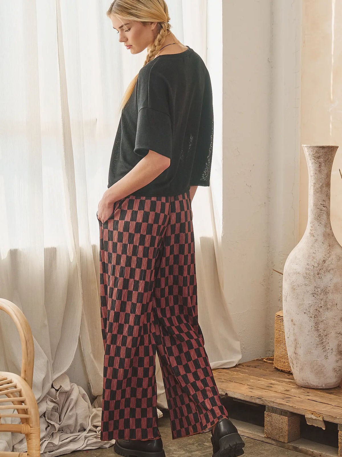A woman stands in a bright room wearing loose black short-sleeve top and wide-leg checkered pants in red and black, with sheer curtains, a wooden bench, and a large vase in the background.