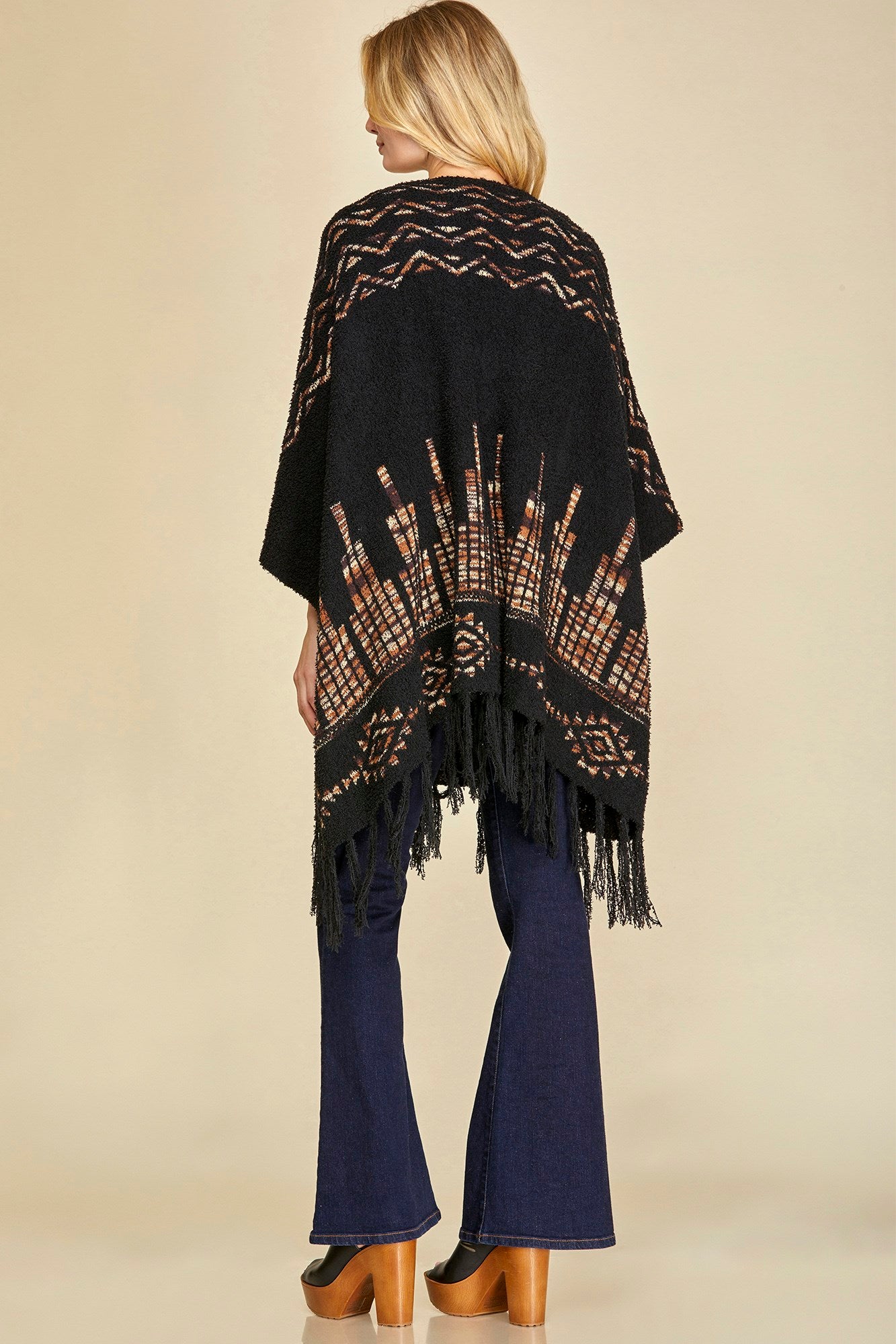 Person wearing a black and brown patterned poncho with fringe on a beige background.