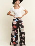 A woman modeling black wide-leg pants with a colorful western-themed patchwork print, paired with a white cropped top and a black cowboy hat, against a solid light beige background.