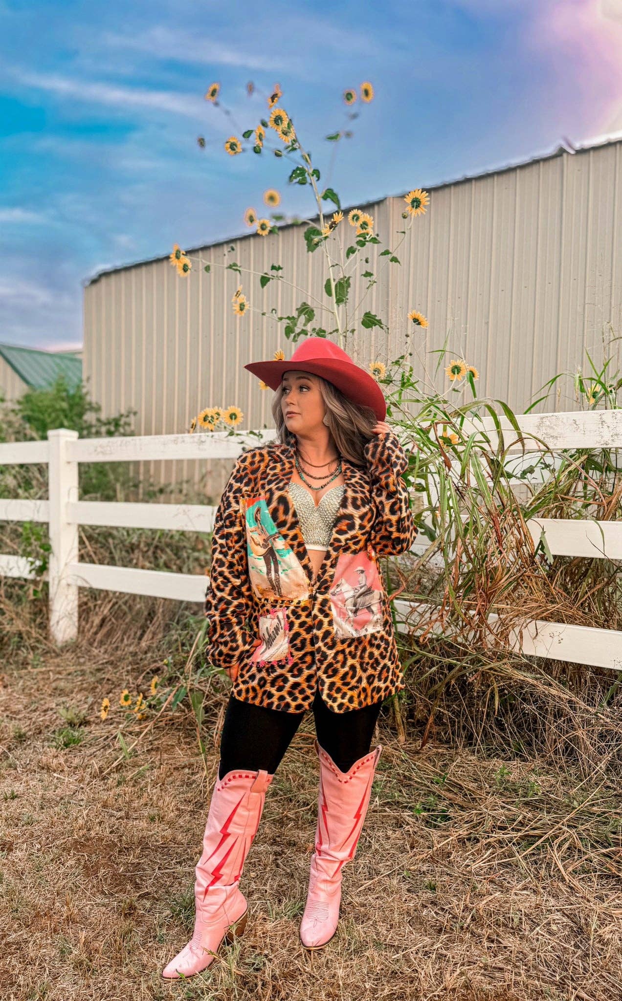 Person wearing a leopard print jacket and pink boots standing in a field with sunflowers and a barn in the background.