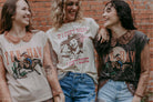 Three women wearing vintage-style t-shirts with cowboy graphics against a brick wall.