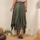 A woman wearing a flowing olive green high-low skirt with a subtle textured pattern, styled with tan cowboy boots in a rustic indoor setting.