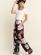A woman modeling a white cropped short-sleeve top and black wide-leg pants with a colorful western-themed graphic print, including a cowboy hat, against a plain beige studio background.