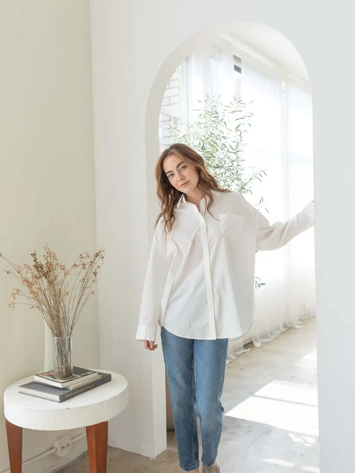 Woman in a white shirt and blue jeans standing in a bright room with a vase of flowers on a small table.