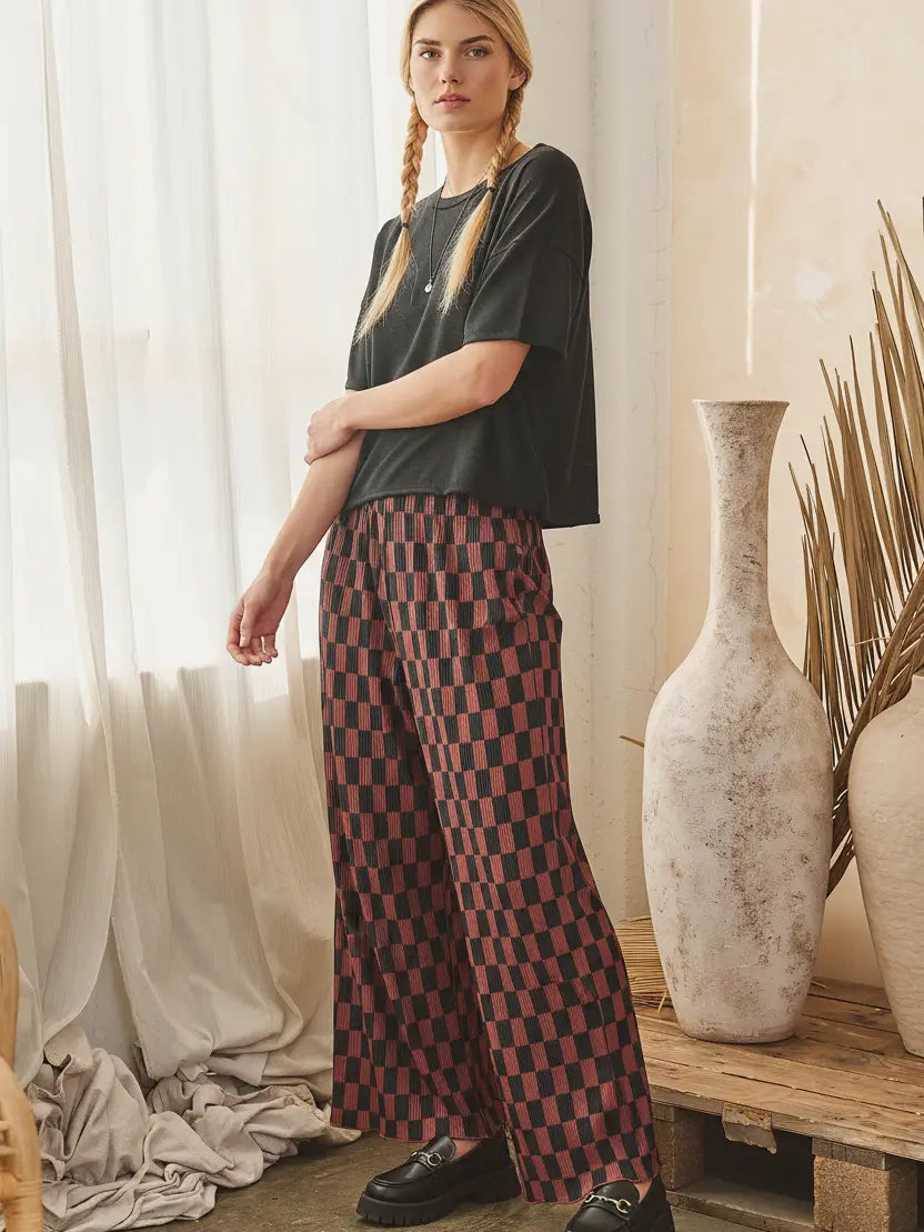 A woman stands indoors wearing a dark charcoal short-sleeve top and wide-leg trousers with a red and dark brown checkerboard pattern, paired with black loafers. The setting features a rustic wooden table, large textured ceramic vases, and sheer white curtains.
