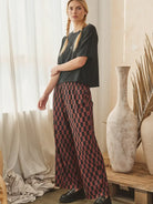 A woman stands indoors wearing a dark charcoal short-sleeve top and wide-leg trousers with a red and dark brown checkerboard pattern, paired with black loafers. The setting features a rustic wooden table, large textured ceramic vases, and sheer white curtains.