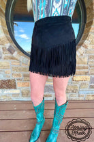 A person wearing a black fringe mini skirt and turquoise western cowboy boots, standing on a wooden deck in front of a stone wall and a round mirror. A 'Sterling Kreek' logo watermark is visible in the bottom right corner.