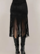 Black fringe skirt worn with black knee-high boots on a plain background