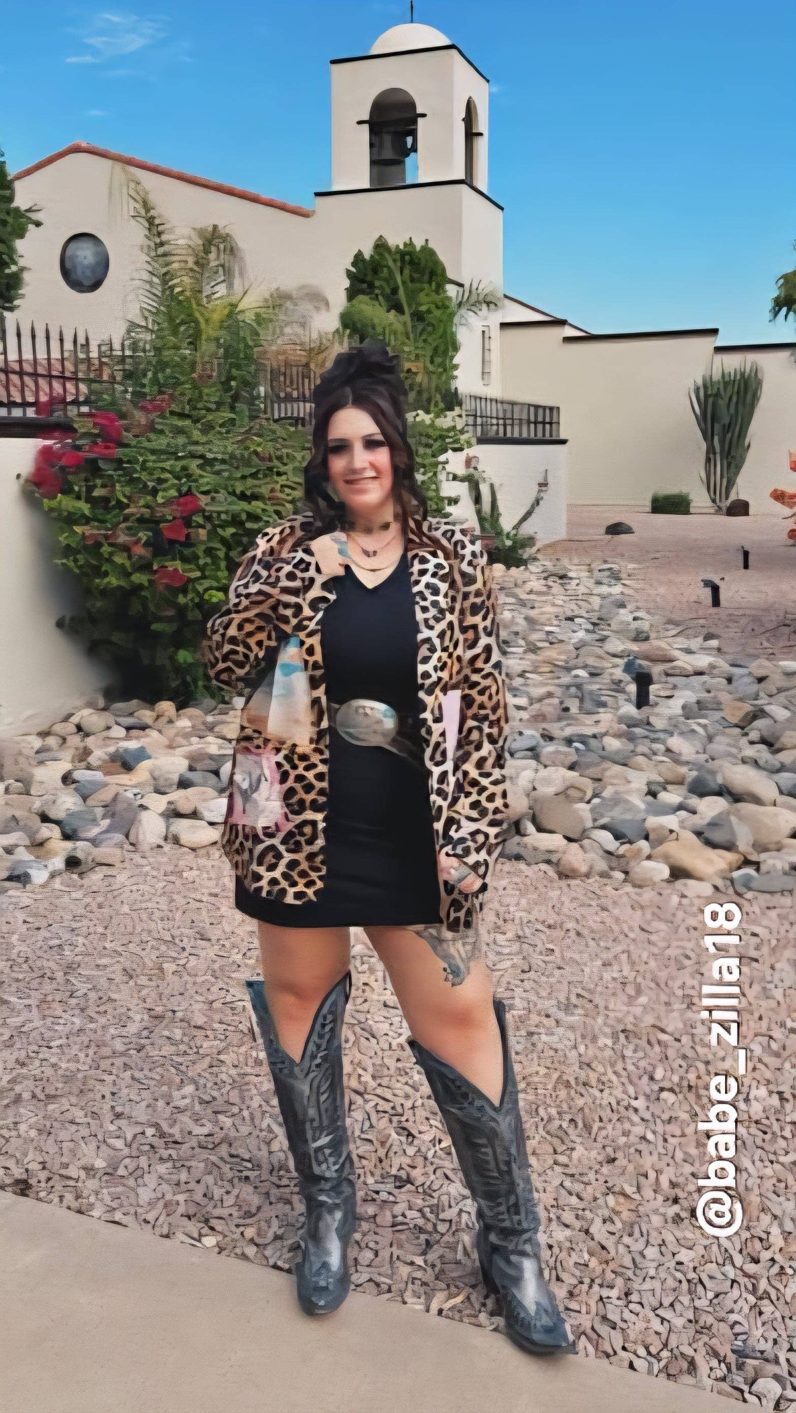 Woman in leopard print cardigan and black dress with cowboy boots standing in front of a building.