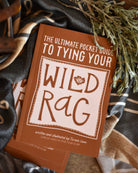 Book titled 'The Ultimate Pocket Guide to Tying Your Wild Rag' on a textured surface with fabric and plants.