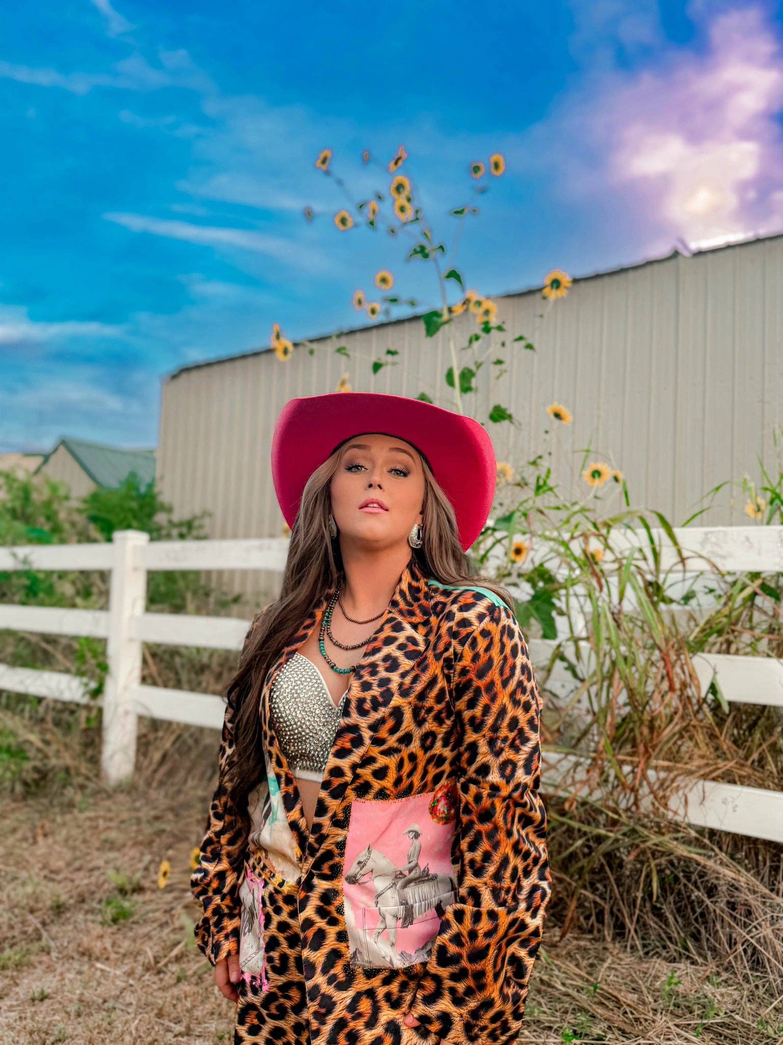 Woman in a leopard print outfit and pink hat standing in a field with sunflowers and a barn in the background.