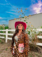 Woman in a leopard print outfit and pink hat standing in a field with sunflowers and a barn in the background.