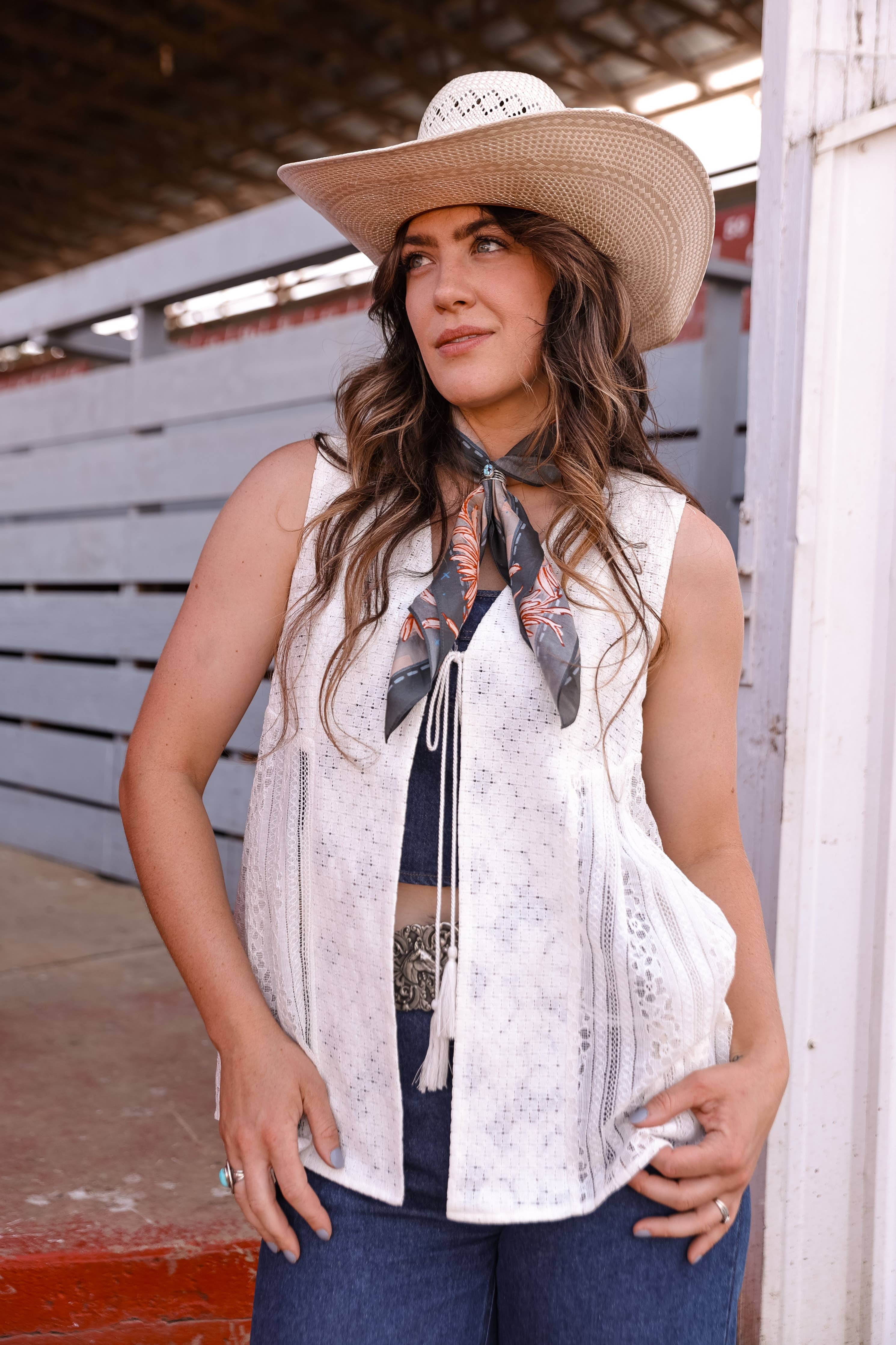 Woman wearing a white sleeveless top and blue jeans with a straw hat and scarf, standing in an indoor setting.