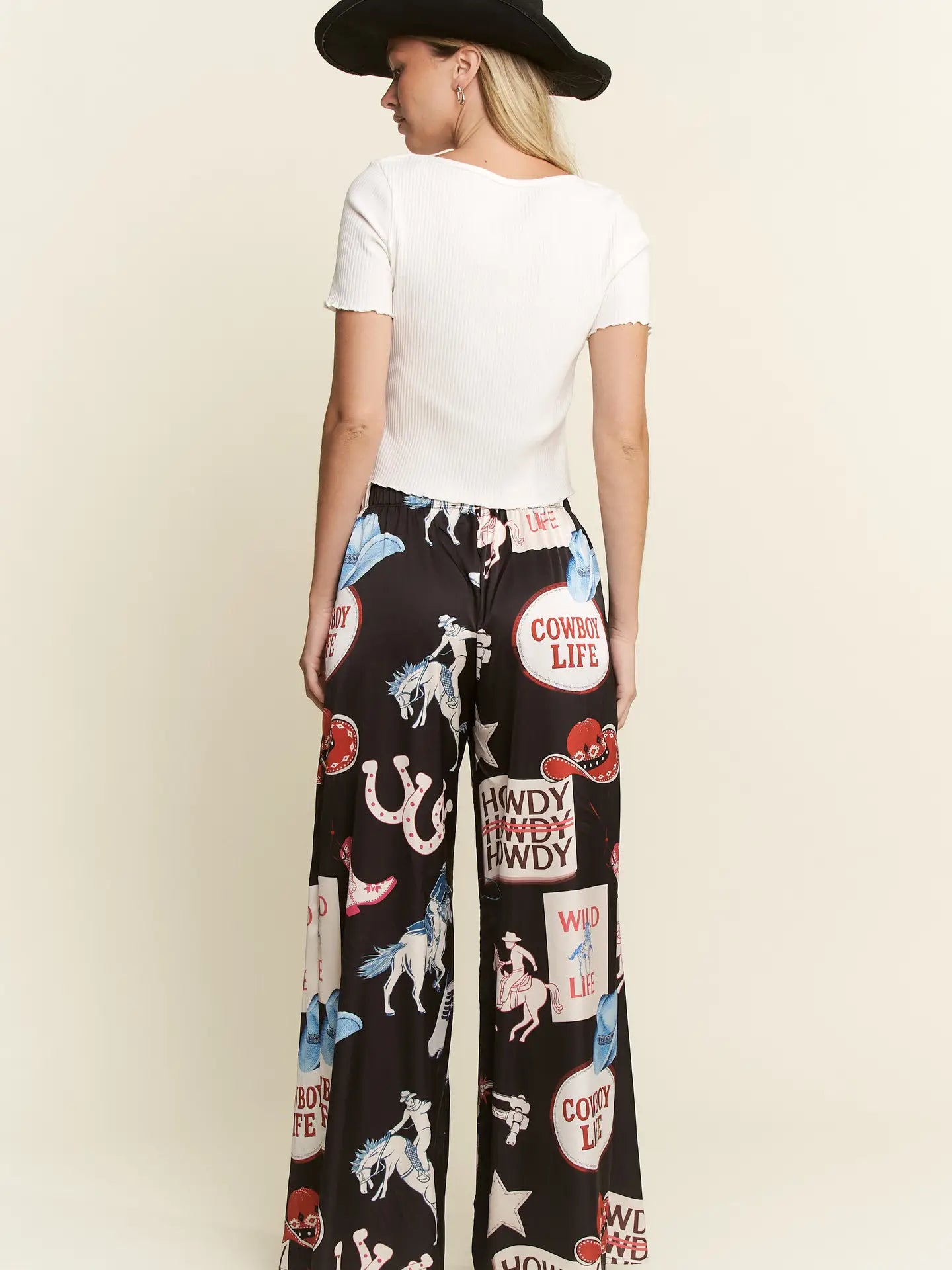 A woman shown from the back wearing wide-leg black pants with a colorful western-themed print (including horses, horseshoes, and cowboy boots) and a white short-sleeve top, standing against a light beige background.