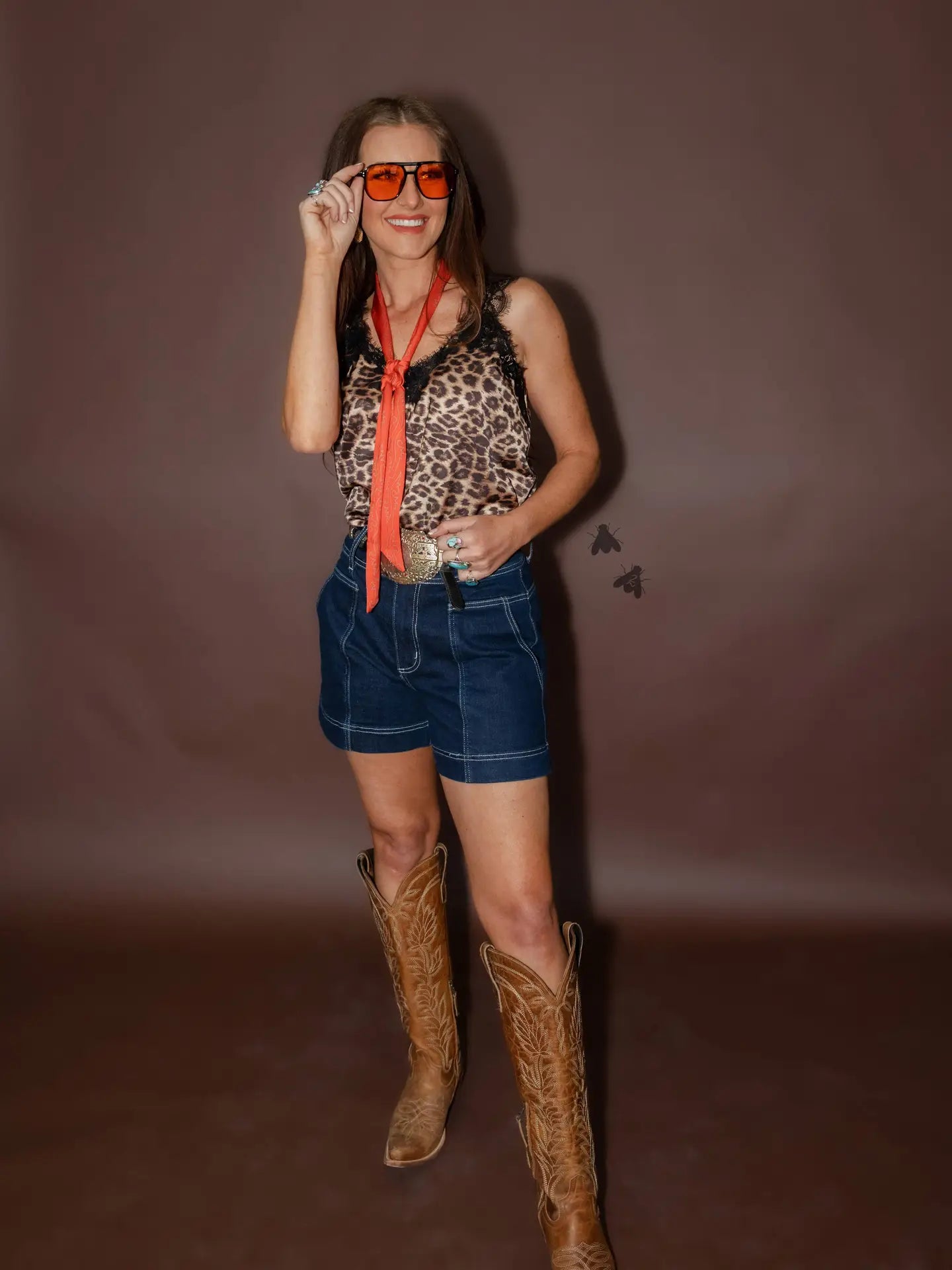 Woman wearing a leopard print top, denim shorts, and cowboy boots against a brown background