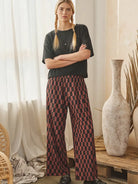 A woman stands indoors with arms crossed, wearing a black short-sleeve top and wide-leg trousers with a red and black checkered pattern. The setting includes sheer curtains, a rustic wooden bench, large ceramic vases, and dried plant stems.