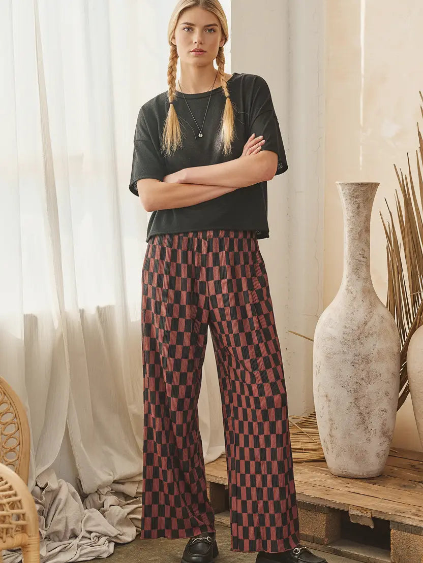 A woman stands indoors with arms crossed, wearing a black short-sleeve top and wide-leg trousers with a red and black checkered pattern. The setting includes sheer curtains, a rustic wooden bench, large ceramic vases, and dried plant stems.