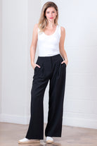 Woman wearing a white sleeveless top and black wide-leg pants against a white brick wall.