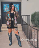 Person wearing a leopard print jacket, black dress, and blue cowboy boots standing outside.