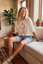 Woman sitting on a couch wearing a beige t-shirt with a logo and denim shorts in a cozy living room.