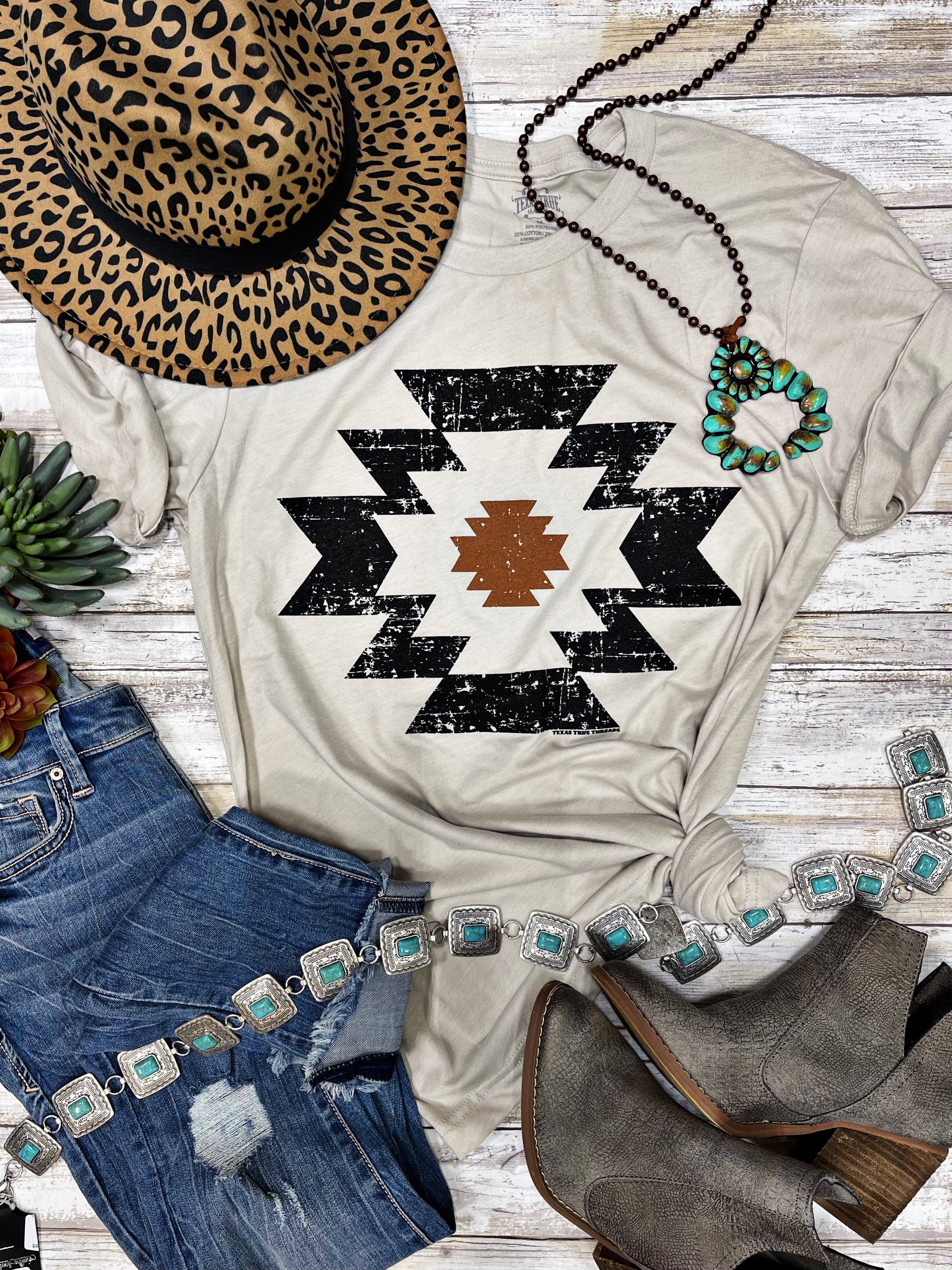 White t-shirt with black geometric pattern, leopard print hat, blue jeans, and brown boots on a wooden surface.