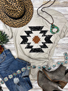 White t-shirt with black geometric pattern, leopard print hat, blue jeans, and brown boots on a wooden surface.