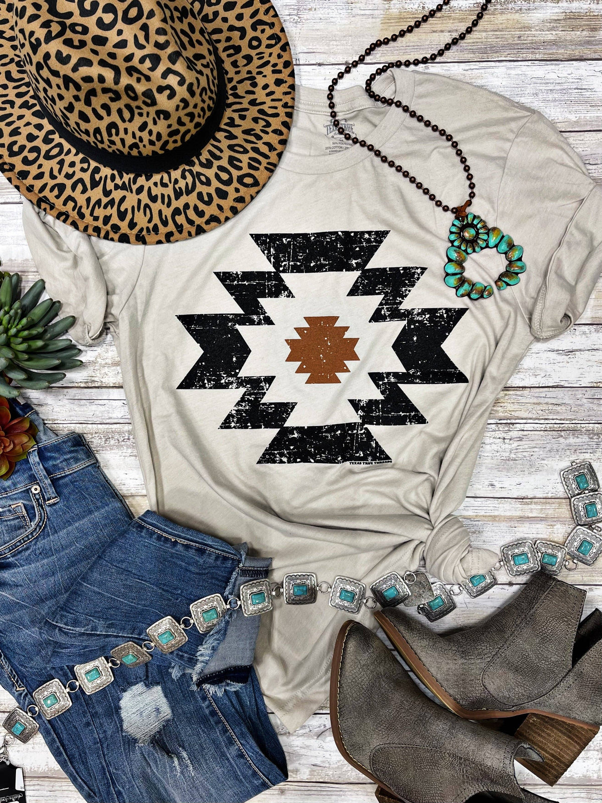 White t-shirt with black geometric pattern, leopard print hat, blue jeans, and brown boots on a wooden surface.