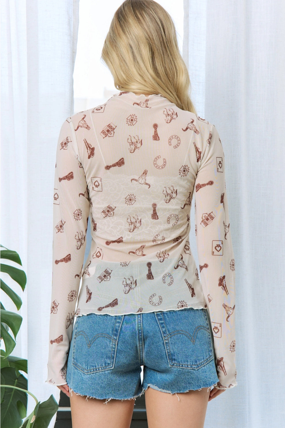 A woman viewed from behind wearing a sheer long-sleeve top with a brown vintage key and lock pattern on a light beige background, paired with blue denim shorts.