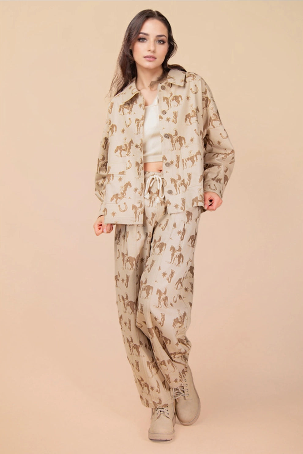 Woman wearing a beige cowboy and horseshoe patterned Shacket  and white hat against a beige background