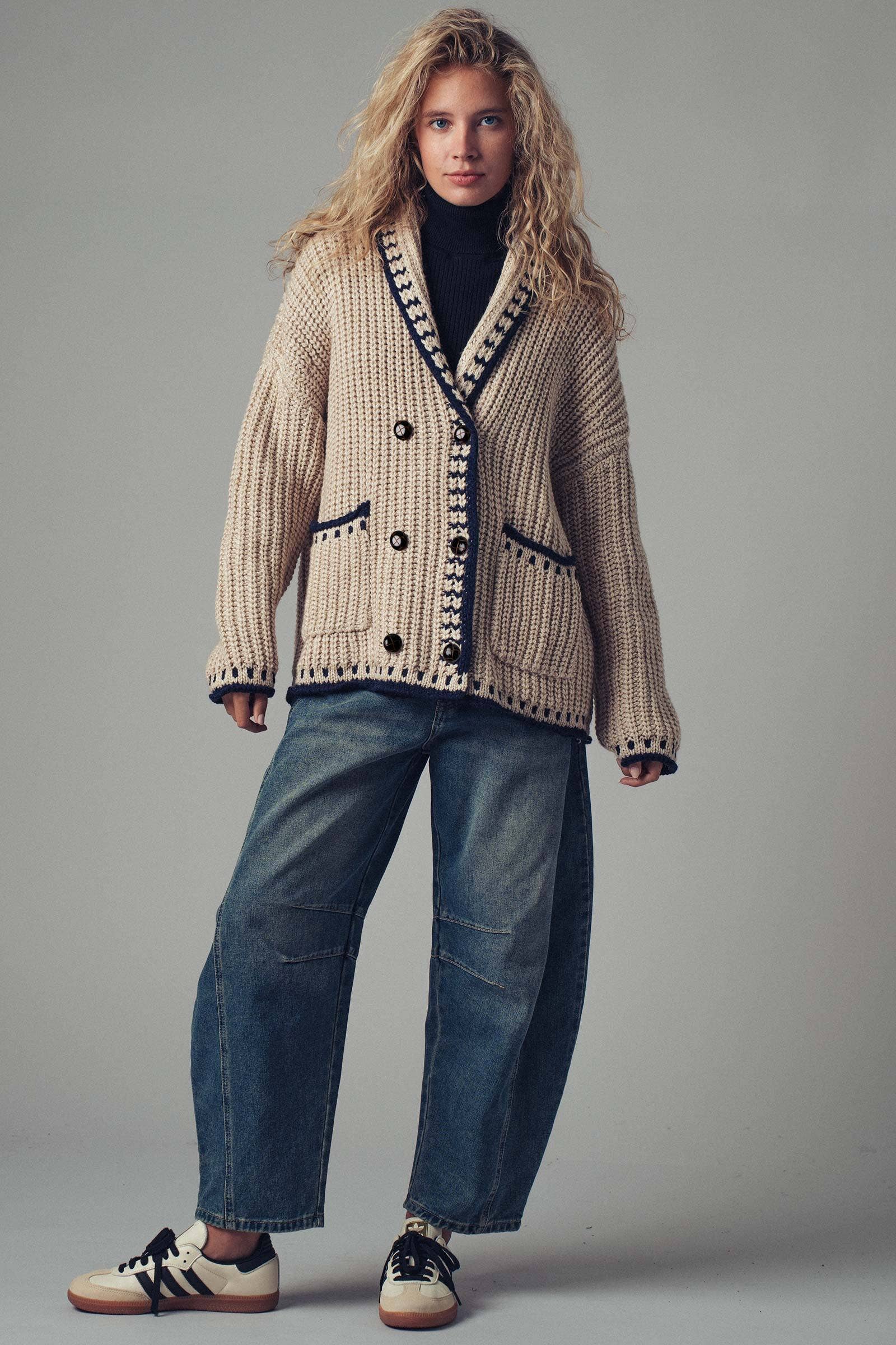 Woman wearing a patterned cardigan, black turtleneck, blue jeans, and sneakers on a gray background