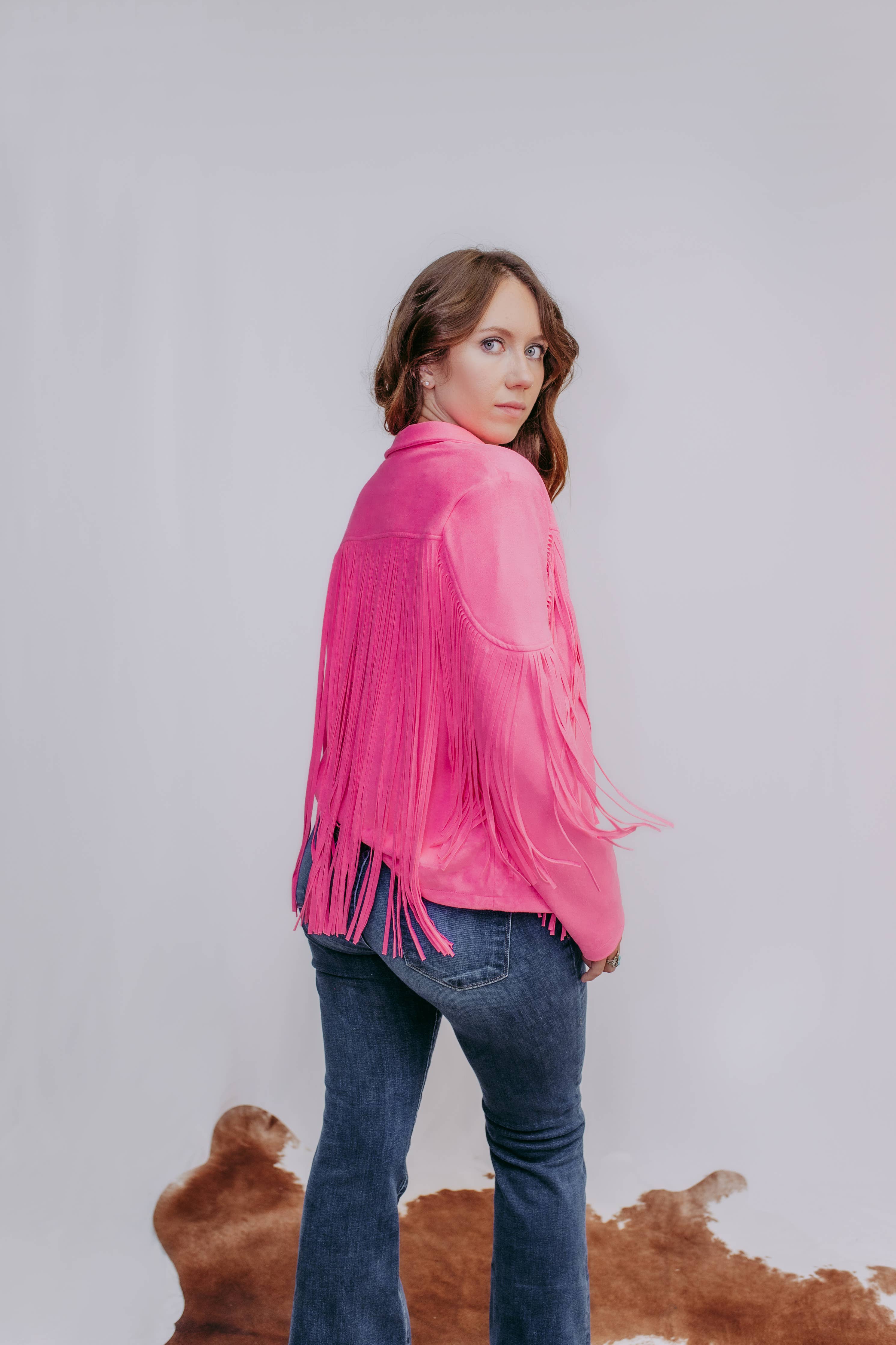 Woman wearing a pink fringed jacket and blue jeans against a plain background