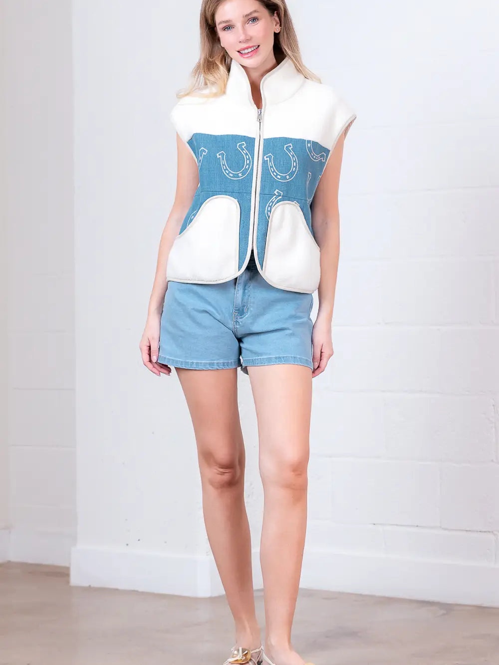 Model wearing indigo denim vest with white horseshoe print and faux-fur collar, zipped front, hands in pockets.”