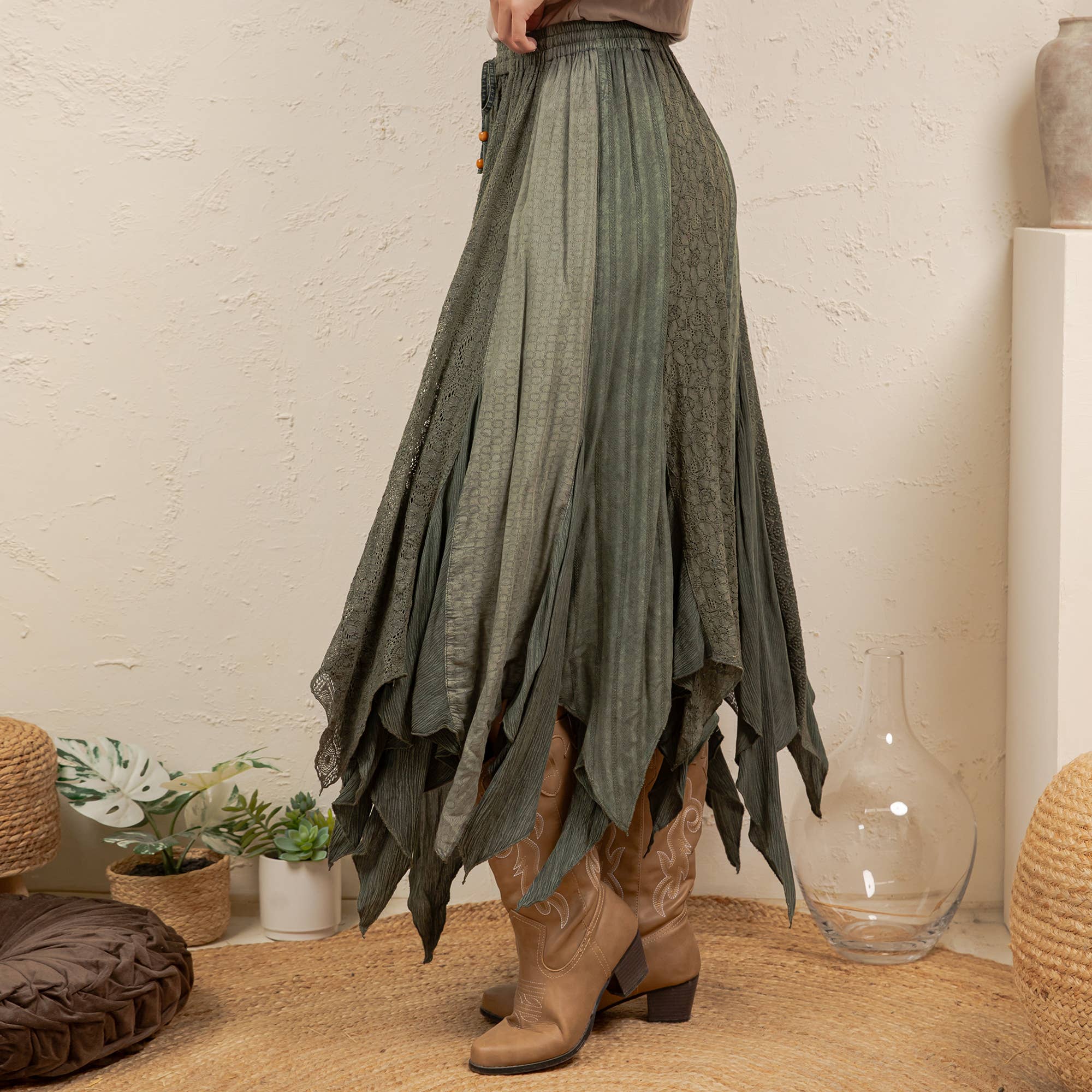 A high-waisted, long pleated skirt in a muted sage green color, styled with brown cowboy boots in a bohemian room setting.