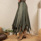 A high-waisted, long pleated skirt in a muted sage green color, styled with brown cowboy boots in a bohemian room setting.