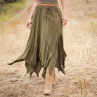 A woman wearing a long, flowing olive green skirt with a tie waist and ruffled asymmetrical hem, walking outdoors in a dry grassy field.